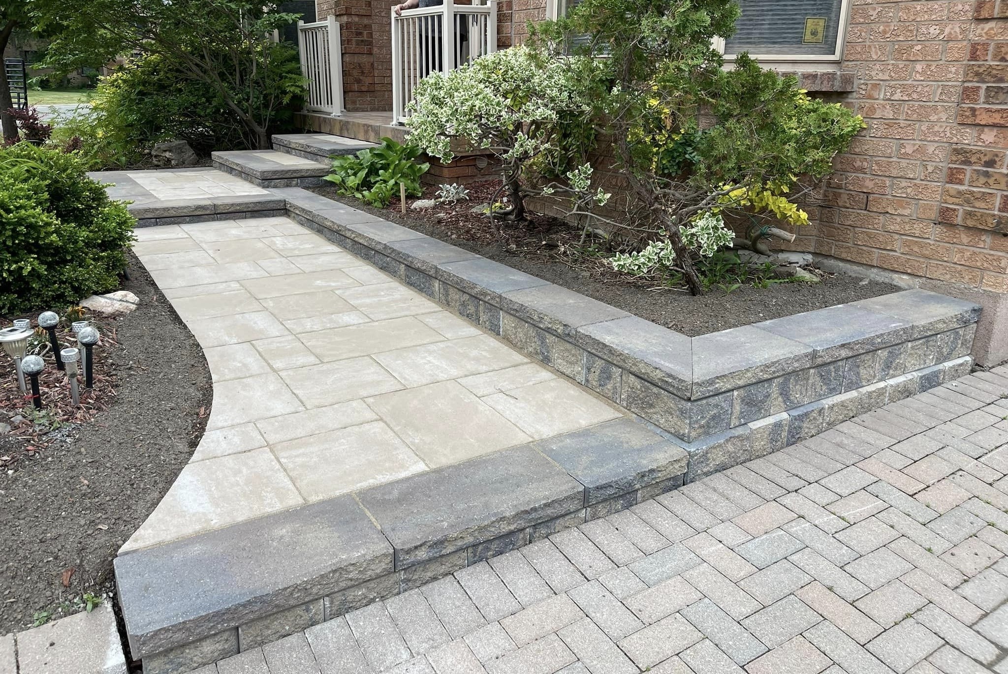 A landscaped walkway with light-colored paving stones and a raised border leading up to a house. Surrounded by plants, lawn, and path lights. Brick facade visible.