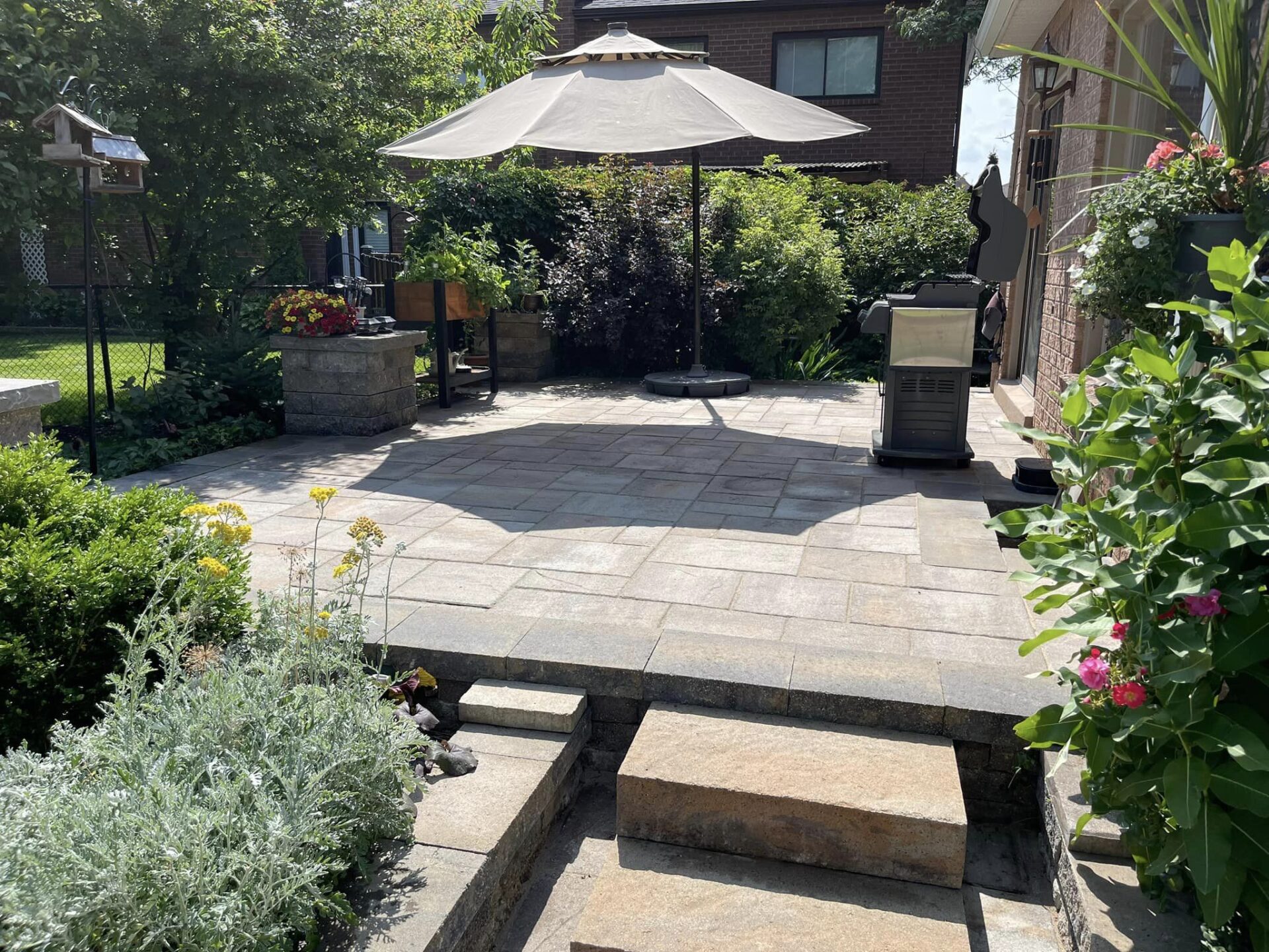 A well-manicured backyard with a stone patio, outdoor heater, large umbrella, greenery, and steps leading up to the paved area under clear skies.