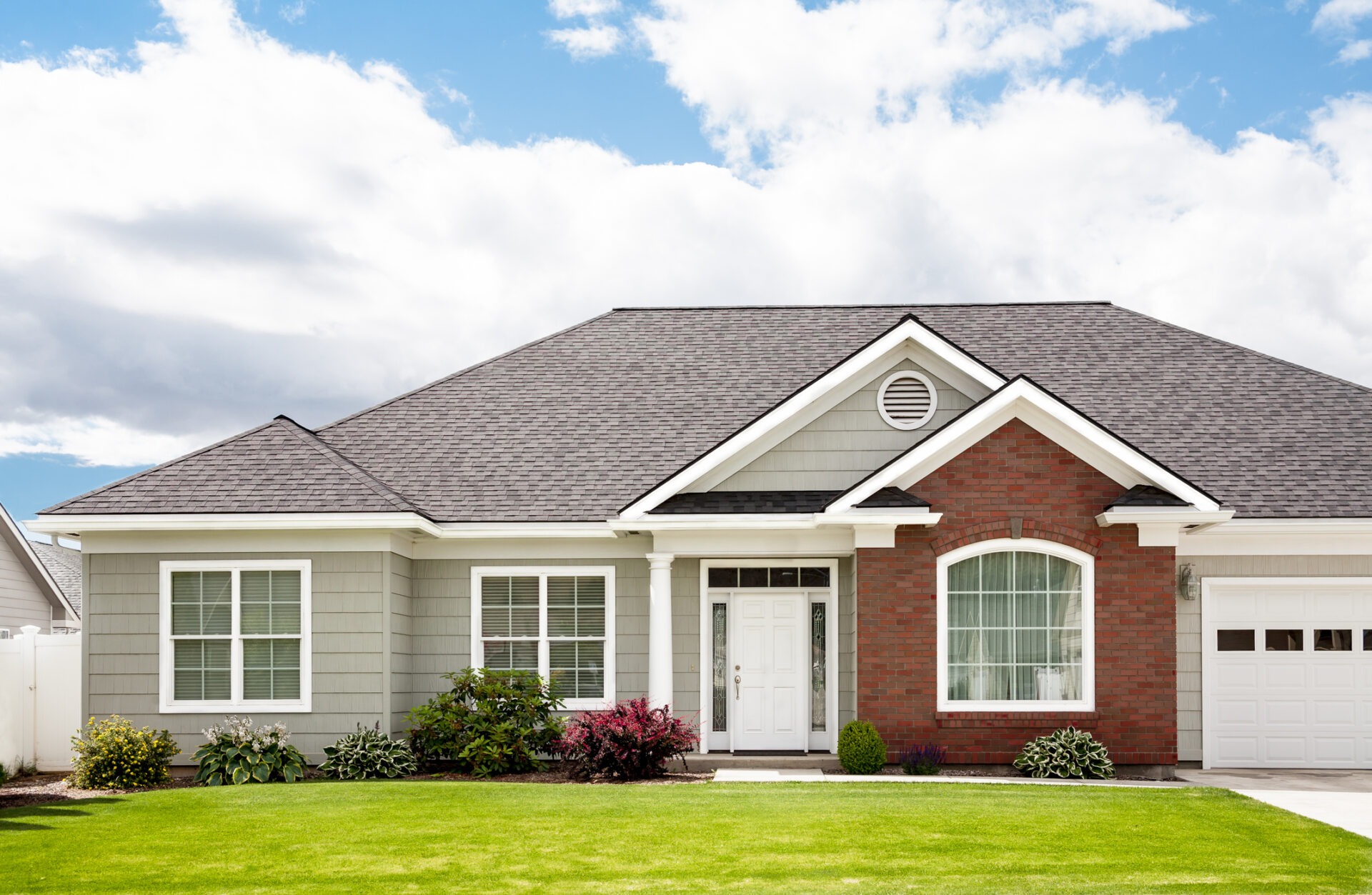 A single-story suburban house with red brick and white siding, a tidy lawn, white garage door, and a background of partly cloudy blue sky.