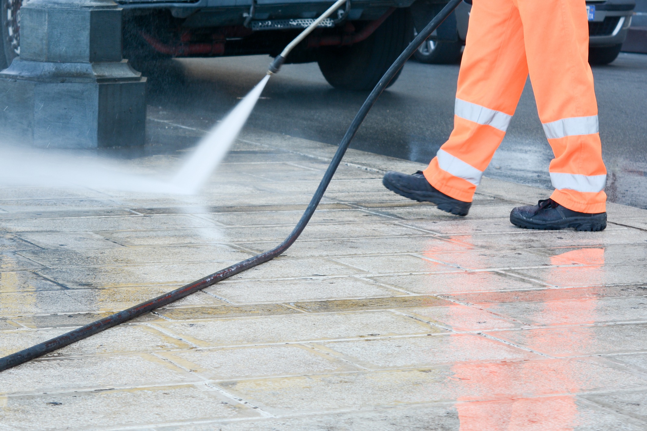 A person in high-visibility orange trousers is pressure washing a dirty urban pavement. Water spray is visible. Part of a vehicle is seen in the background.