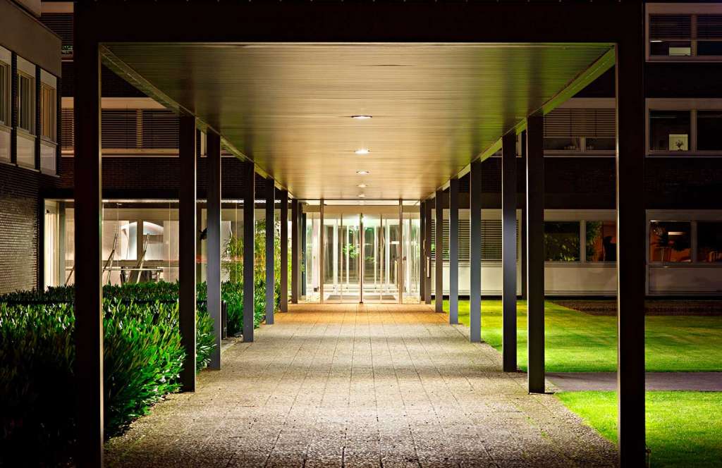 A well-lit, modern walkway with a metal roof structure, paved floor, leading to glass doors. Flanked by buildings and landscaped grass at night.