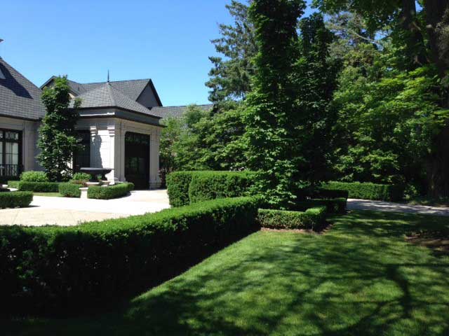 This is an image of a manicured garden in front of an elegant house with a well-kept lawn, hedges, and mature trees under a clear blue sky.