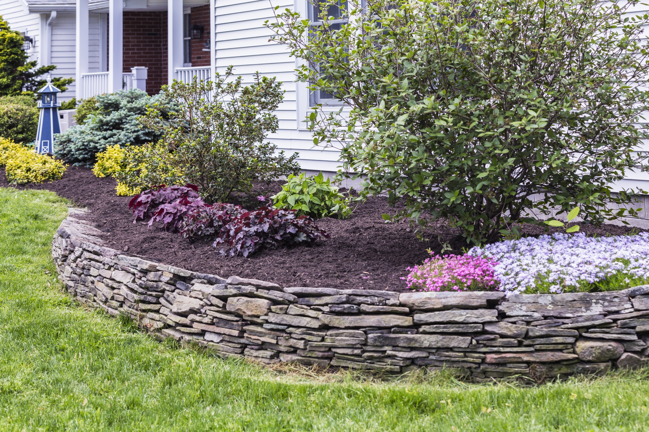 A well-maintained garden bed with a stone wall border in front of a traditional house. Various plants and blooming flowers create a colorful landscape scene.