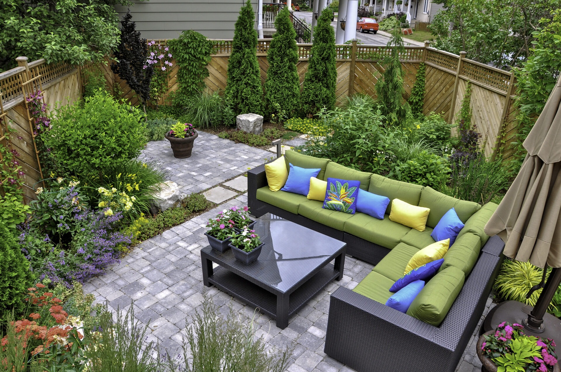 This image shows a cozy garden patio with a green outdoor couch adorned with colorful pillows, a stone pathway, lush plants, and a wooden fence.