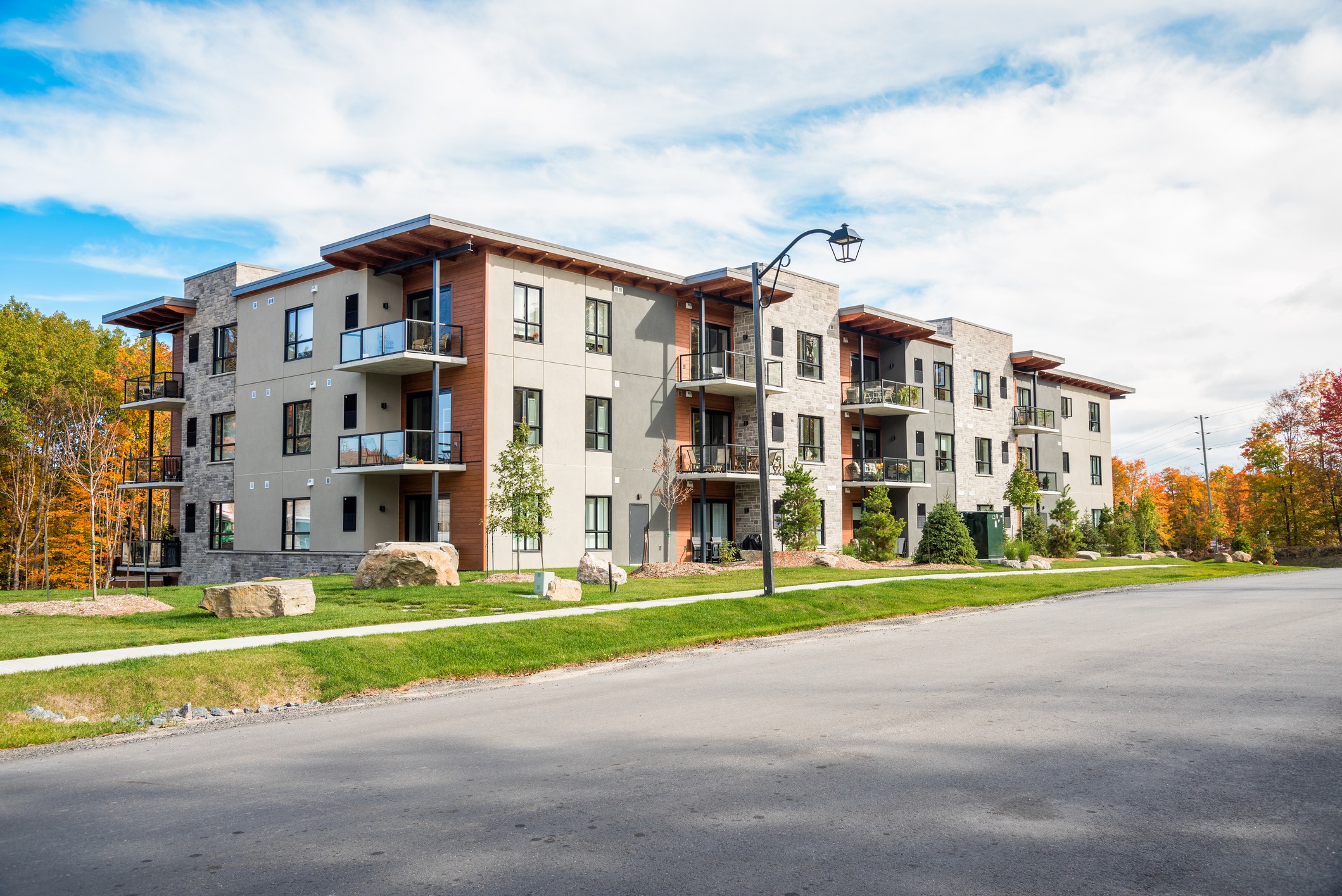 A modern, multi-story residential building with balconies, large windows, and a mix of stone and stucco exteriors, set against a backdrop of autumn foliage.