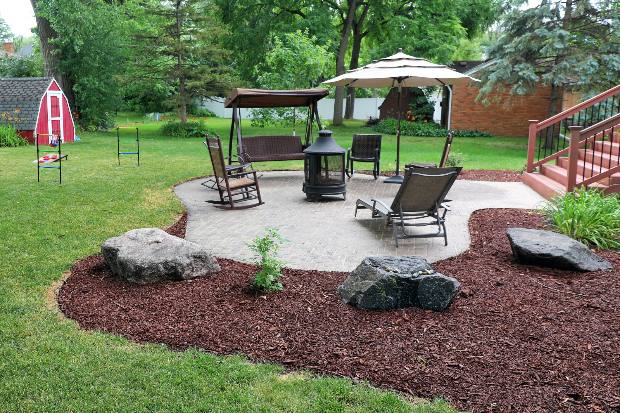 This image depicts a landscaped backyard with a swing seat, chairs, an umbrella, and a gazebo on a brick patio. There are large rocks and a small shed.