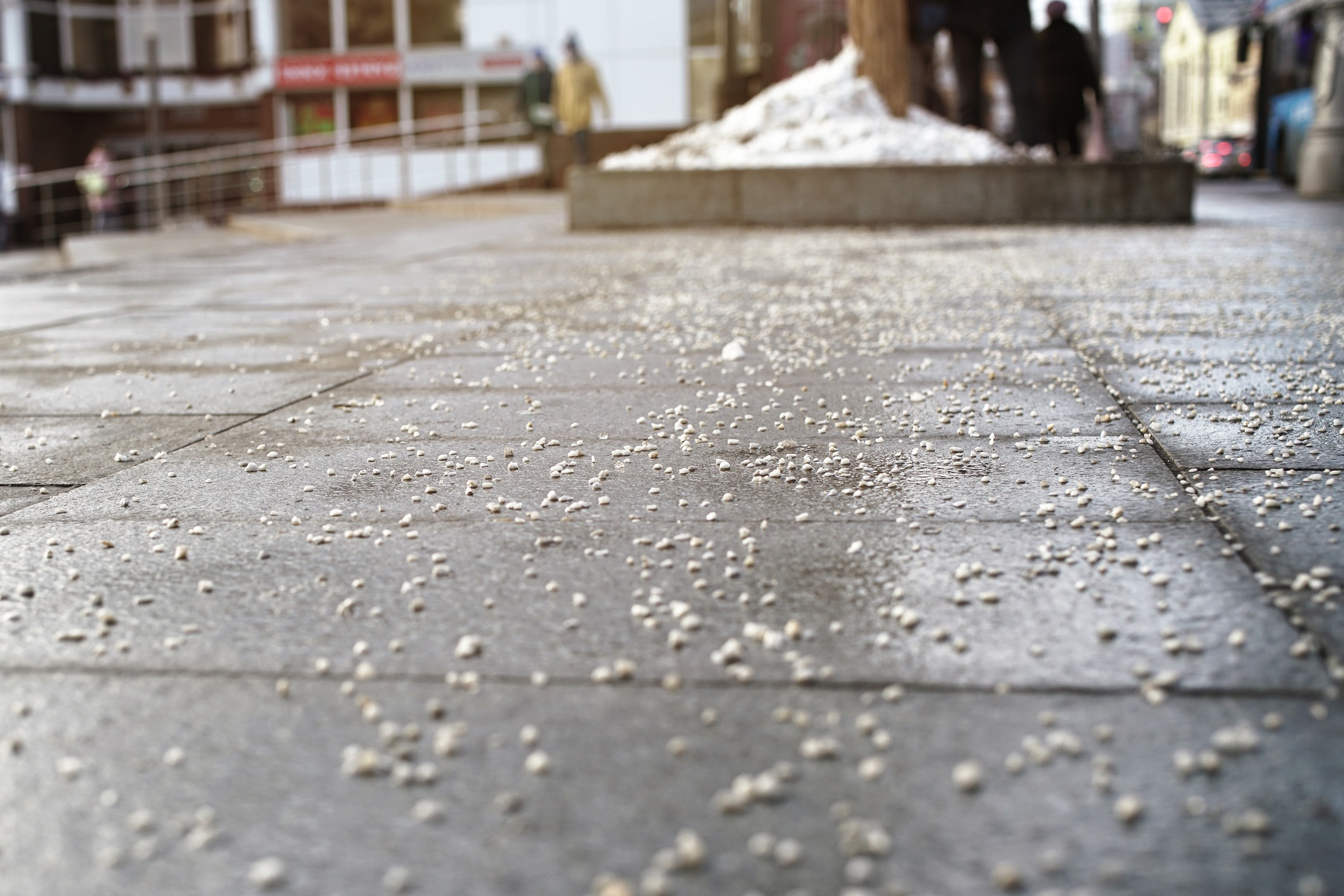 A snowy walkway with scattered salt. People walk in the background. Buildings and a tree planter are visible, suggesting an urban winter scene.