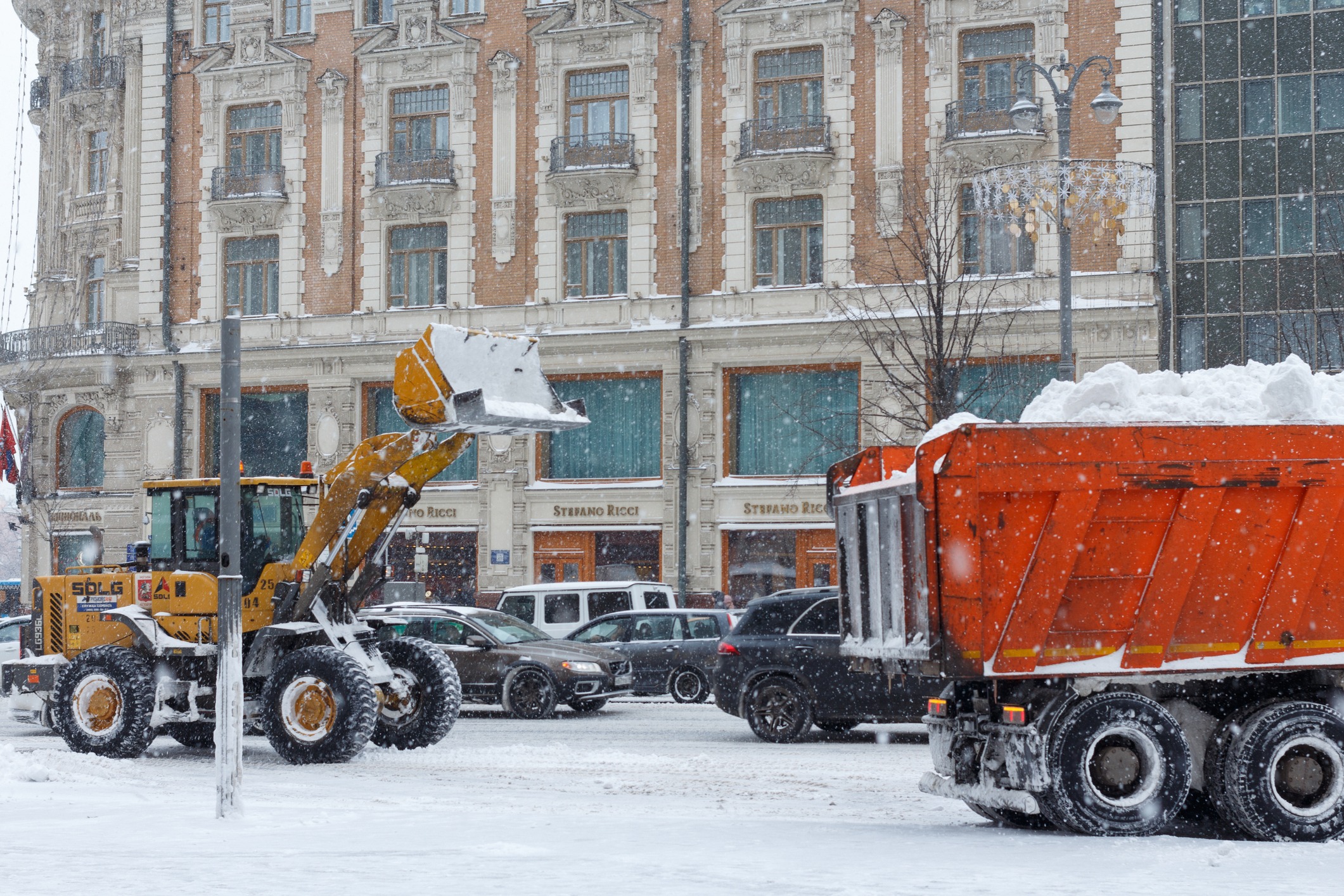 A snow-covered street with vehicles, a bulldozer, and a snow truck in front of the historic Metropole Hotel, Moscow, during snowfall.