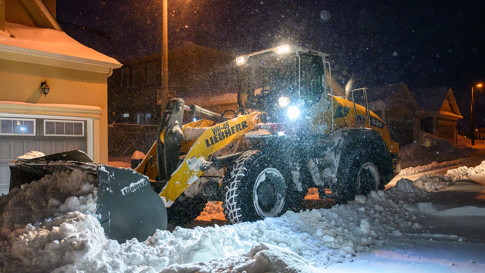 A snowplow clears snow at night on a residential street; streetlights illuminate the scene, showing snow-covered houses and vehicles.