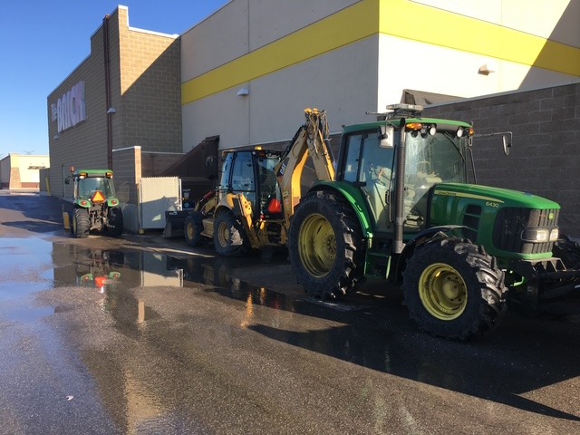 Three tractors are parked outside a large store building on a sunny day, with wet pavement reflecting sunlight.