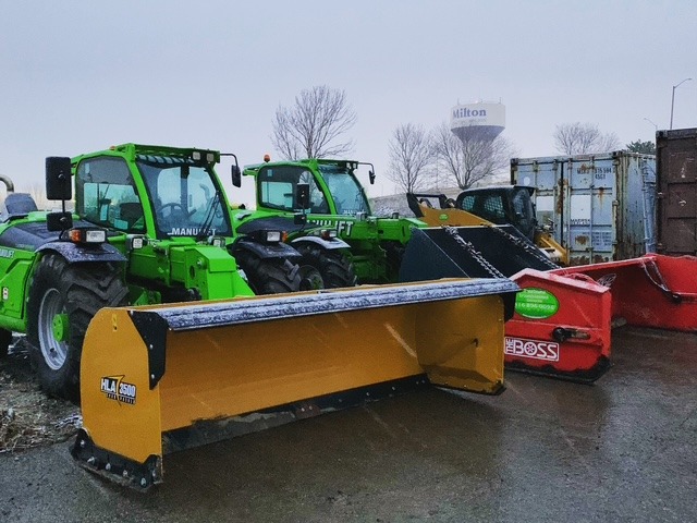 Green construction vehicles with snowplows are parked on a cloudy day. The Hilton water tower is visible in the background.
