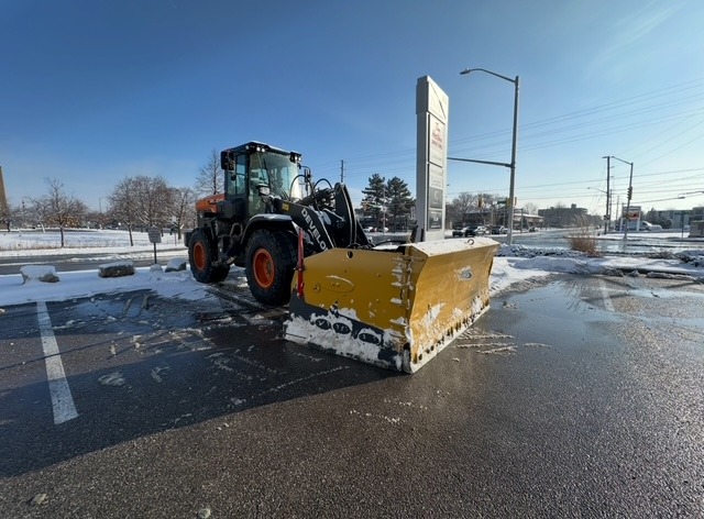 A snowplow vehicle clears snowy parking lot under clear blue sky. Trees and utility poles line the background; no landmarks are visible.