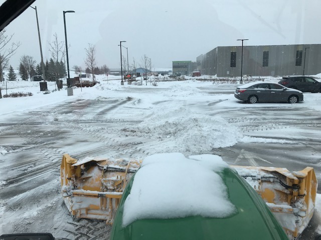 A snowplow clearing a snow-covered parking lot. Cars parked nearby, buildings in the background under overcast sky. No recognizable landmarks visible.